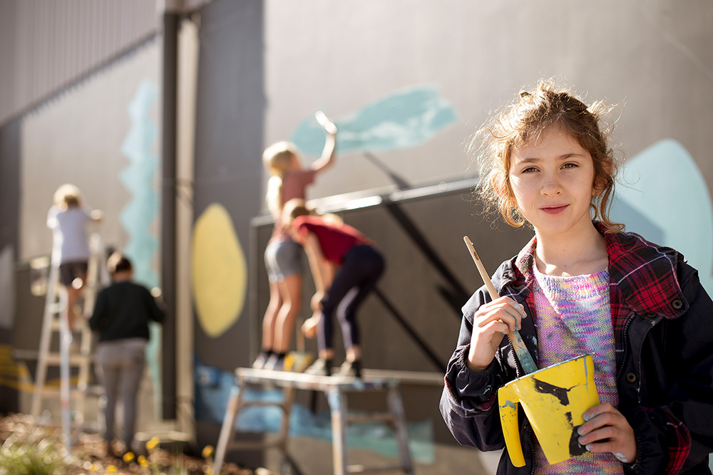 A girl standing with a bucket of paint holding a paint brush looking into the camera in front of a mural being painted by other people.
