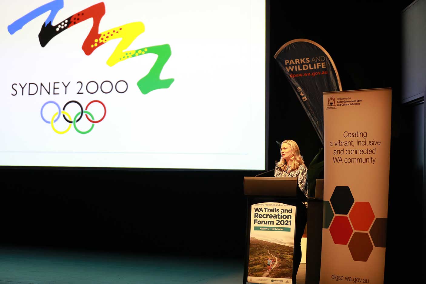 A person speaking at a lectern on stage. A Sydney 2000 Olympic logo is on screen.