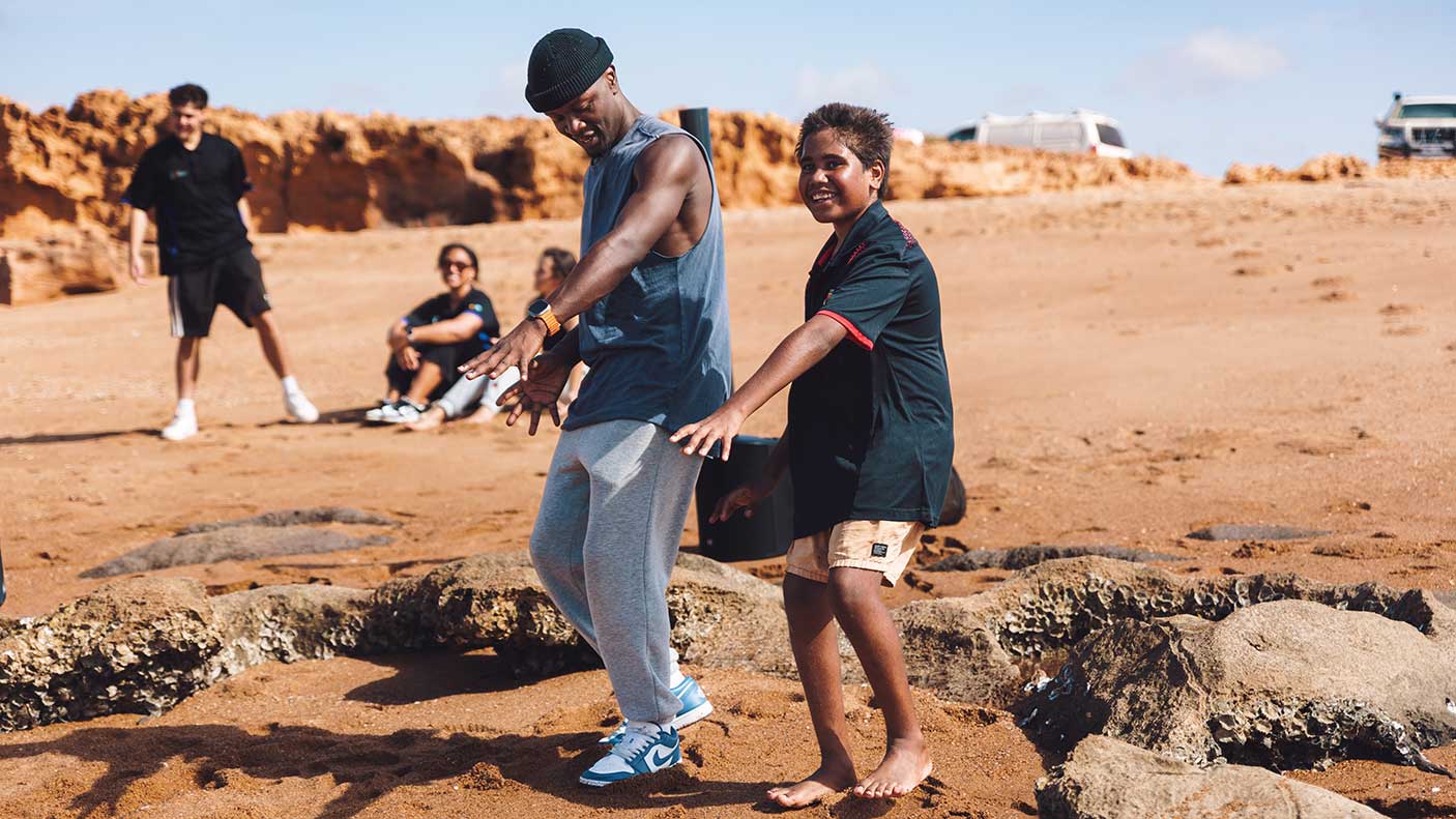 A man and a boy posing a dance move in a desert environment. A group of people are watching in the background.
