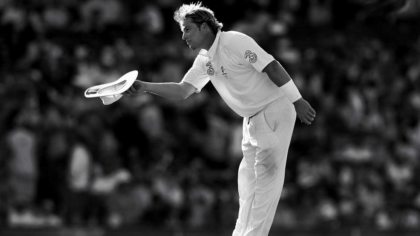 Black and white photograph of Shane Warne holding his hat and bowing on a cricket field