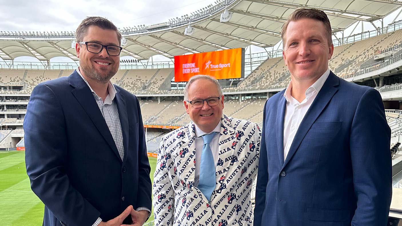 3 men wearing suits at Optus Stadium with the True Sport branding on the big screen behind them.